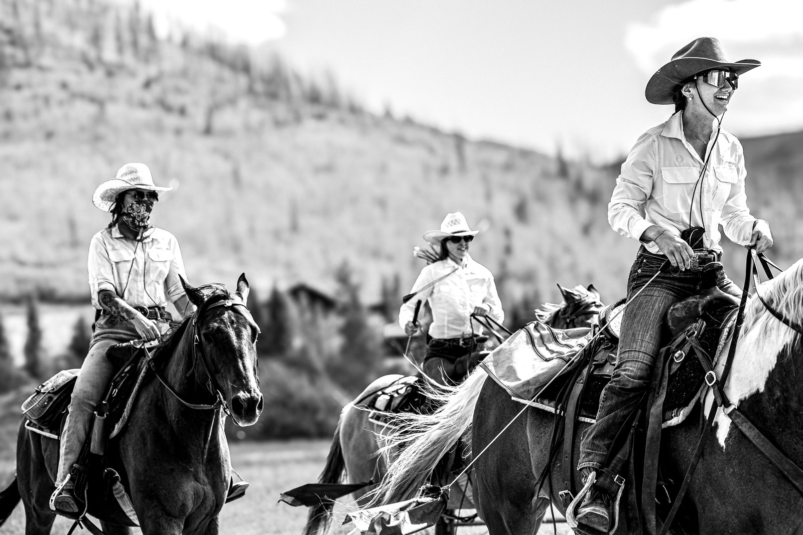 Three cowgirls in western hats ride horses across a sunlit valley, the foreground rider laughing behind reflective sunglasses in high-contrast black and white.