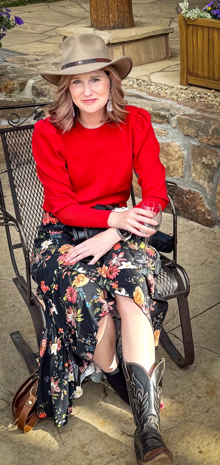 The photographer, seated on a ranch stone patio in a wide-brimmed hat and cowboy boots, camera resting in her lap and a glass of wine in hand.