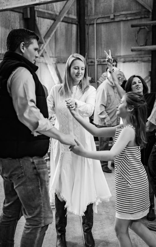 Black-and-white barn scene of a man, a woman in a white dress and cowboy boots, and a girl in a striped dress dancing.