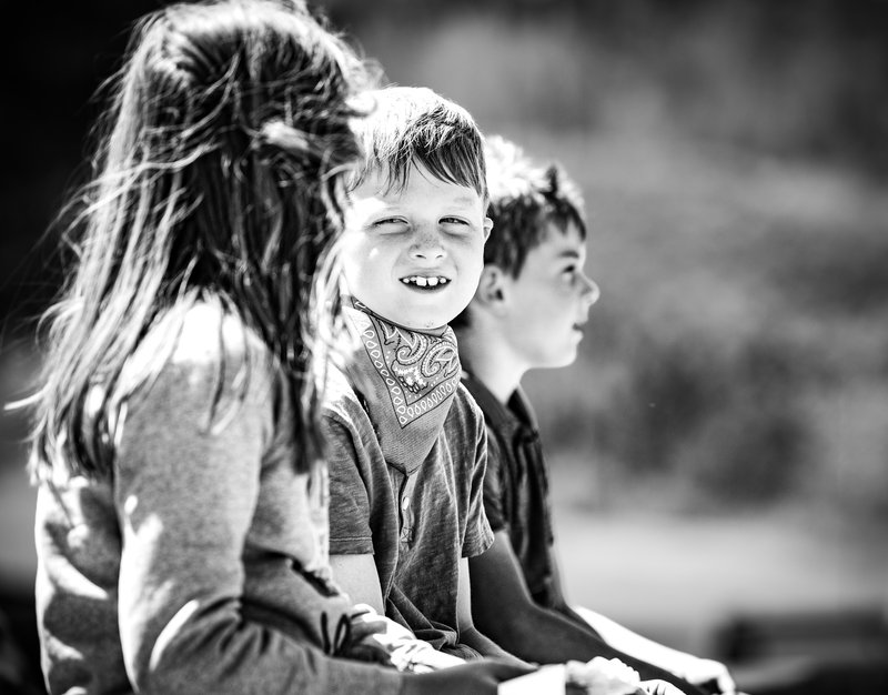 A gap-toothed boy in a bandana grins directly at the camera between a foreground girl and another boy, in soft light.