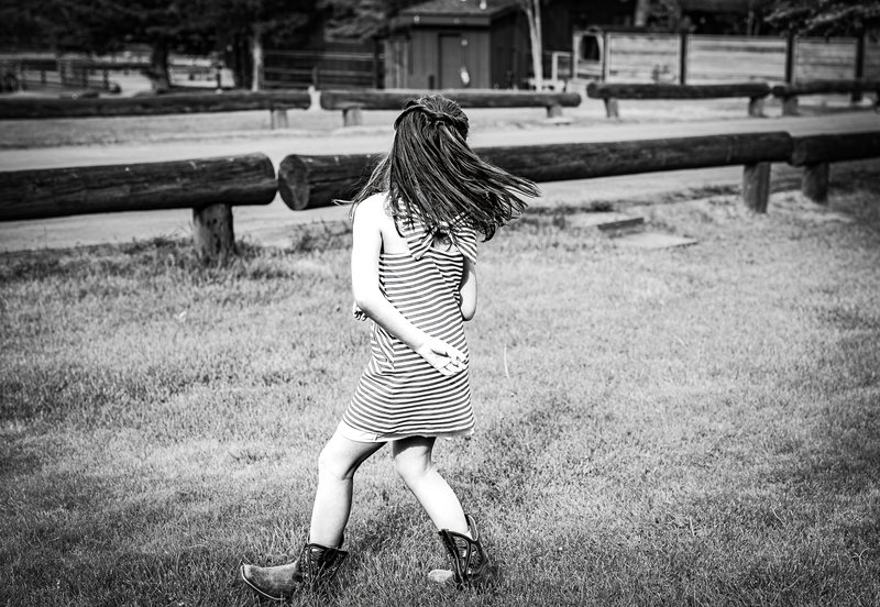 A young girl in a striped dress and cowboy boots spins on a grassy field, her long hair whipping across her face.