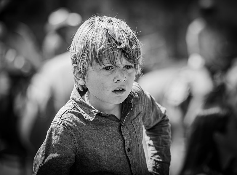 Black-and-white portrait of a tousle-haired young boy with a furrowed, intense expression against a blurred crowd.