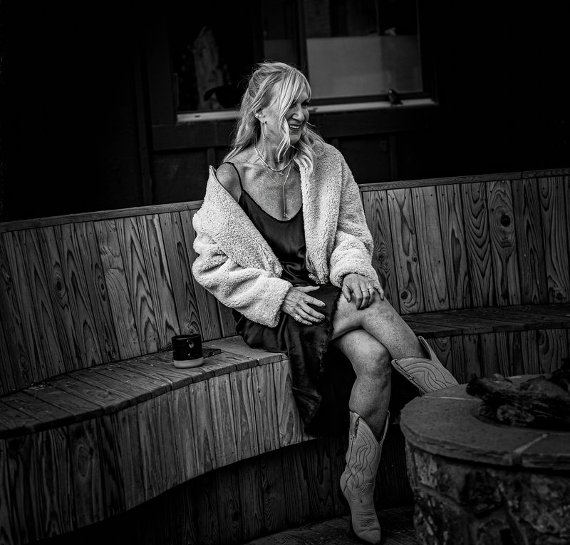 A laughing blonde woman in a shearling coat and cowboy boots sits on a curved wooden bench, gazing off-camera.
